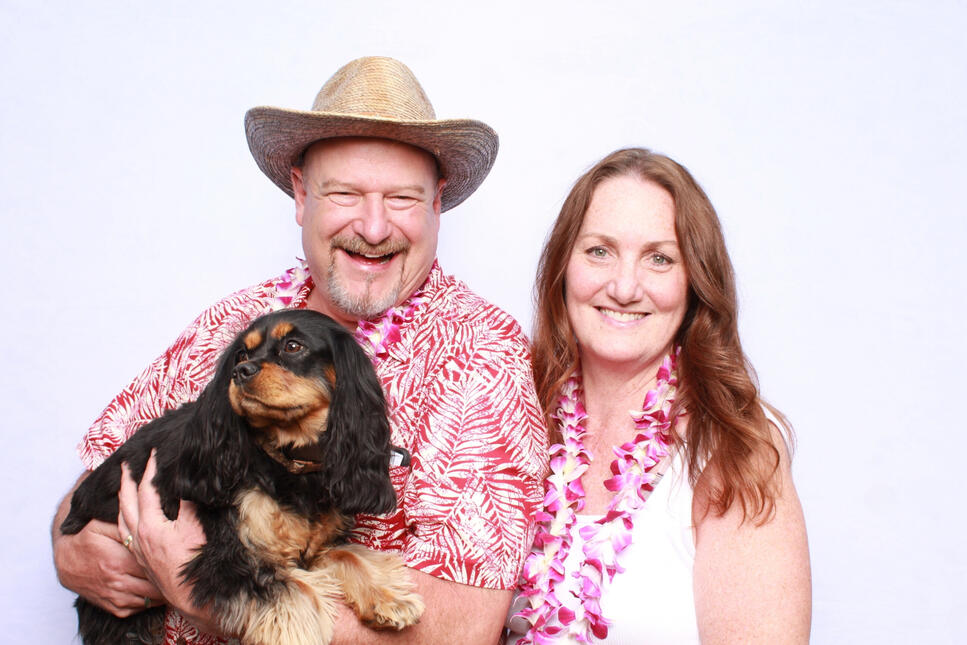 Couple smiling for a photo booth photo wearing Hawaiian shirts and leis, man wearing a straw hat, holding a Cavalier King Charles Spaniel